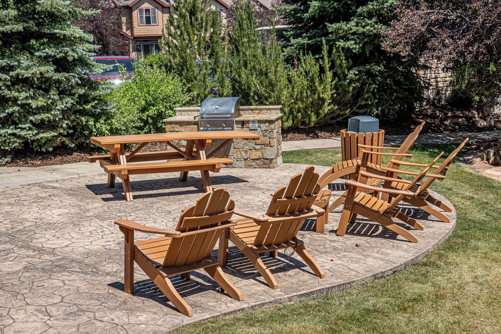 An outdoor patio with a stone table and wooden chairs, a barbecue grill, and a stone fireplace.