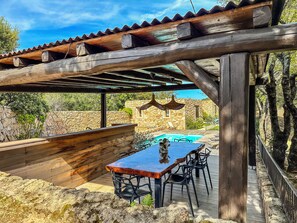 Eine hölzerne Pergola mit Poolbillard und Stühlen darunter, umgeben von Steinmauern und Grünpflanzen.