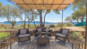 Un espace extérieur aménagé, abrité et doté de meubles en osier, avec une table préparée pour deux personnes, offrant une vue panoramique sur un paysage verdoyant, avec des arbres et des montagnes à l’horizon.