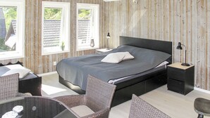 Un dormitorio con una cama grande, dos ventanas, una pared de madera, una mesita y una silla.