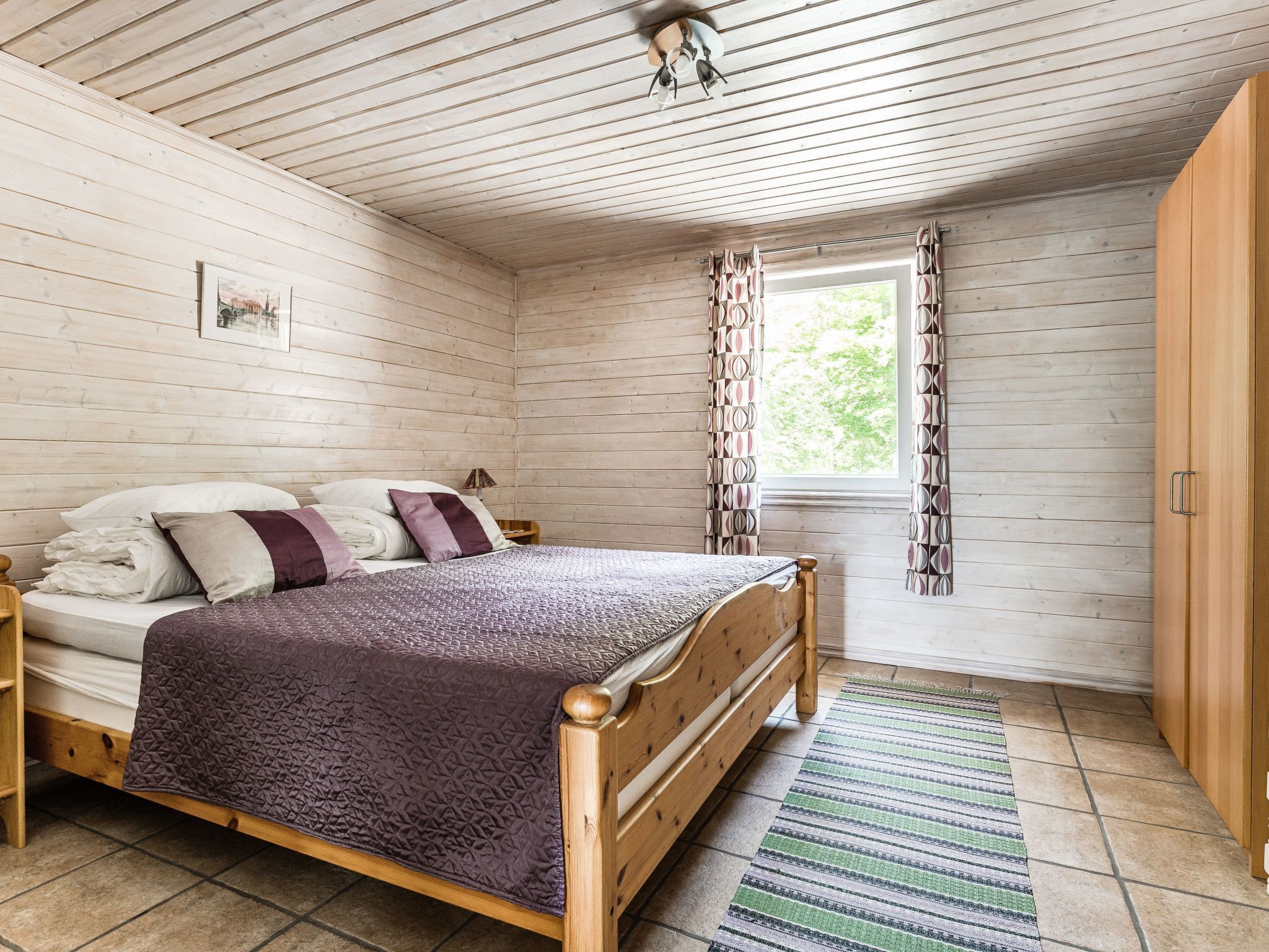 Une chambre à coucher aux murs lambrissés, comprenant un lit, une table de chevet, une fenêtre avec des rideaux et une armoire en bois.