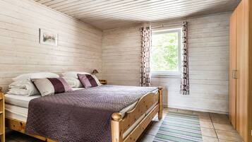 Une chambre à coucher aux murs lambrissés, comprenant un lit, une table de chevet, une fenêtre avec des rideaux et une armoire en bois.