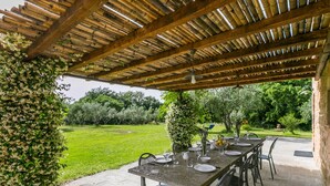 Une terrasse extérieure avec une longue table dressée pour le repas, entourée de chaises, sous une pergola en bois offrant une vue sur un jardin verdoyant.