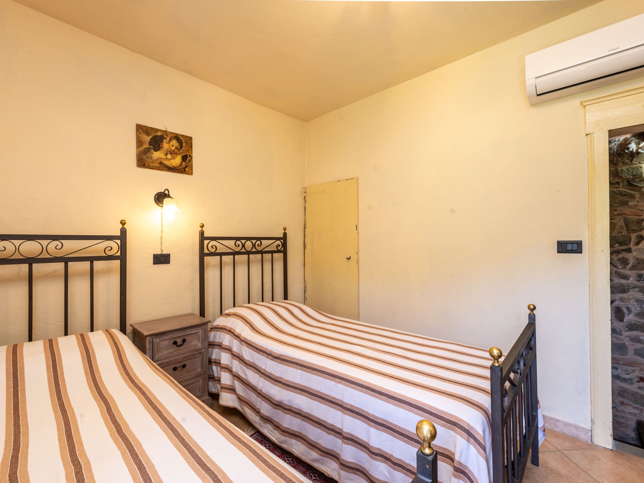 A bedroom with two beds, a nightstand, a wall-mounted lamp, a painting, and an air conditioning unit.