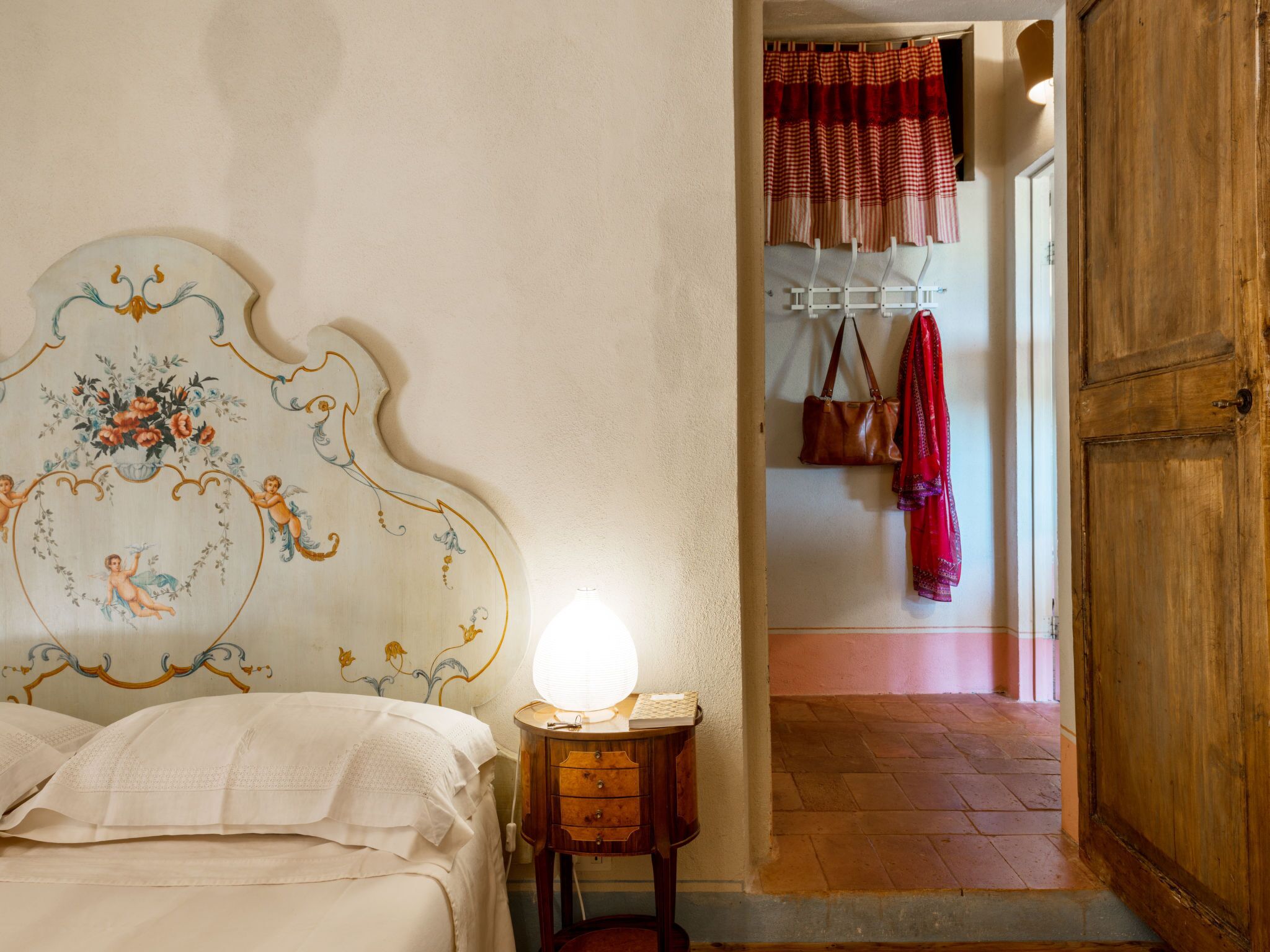 Un dormitorio con cama, mesita de noche, lámpara, puerta de madera y un pasillo que conduce a una habitación con cortina a cuadros rojos y perchero.