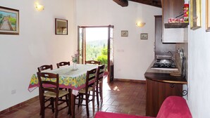 Un coin repas avec une table et des chaises, une cuisine équipée d’une cuisinière, et une vue sur la verdure par une porte ouverte.