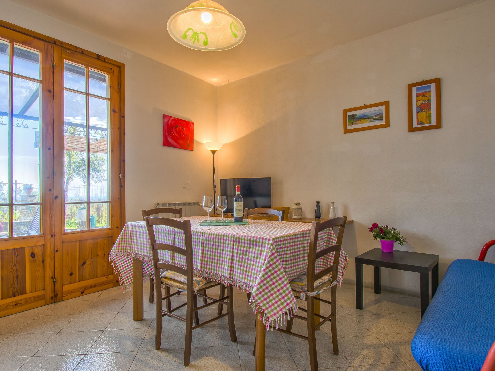 A dining area with a table set for two, wooden chairs, a television, and a window with a view of greenery.