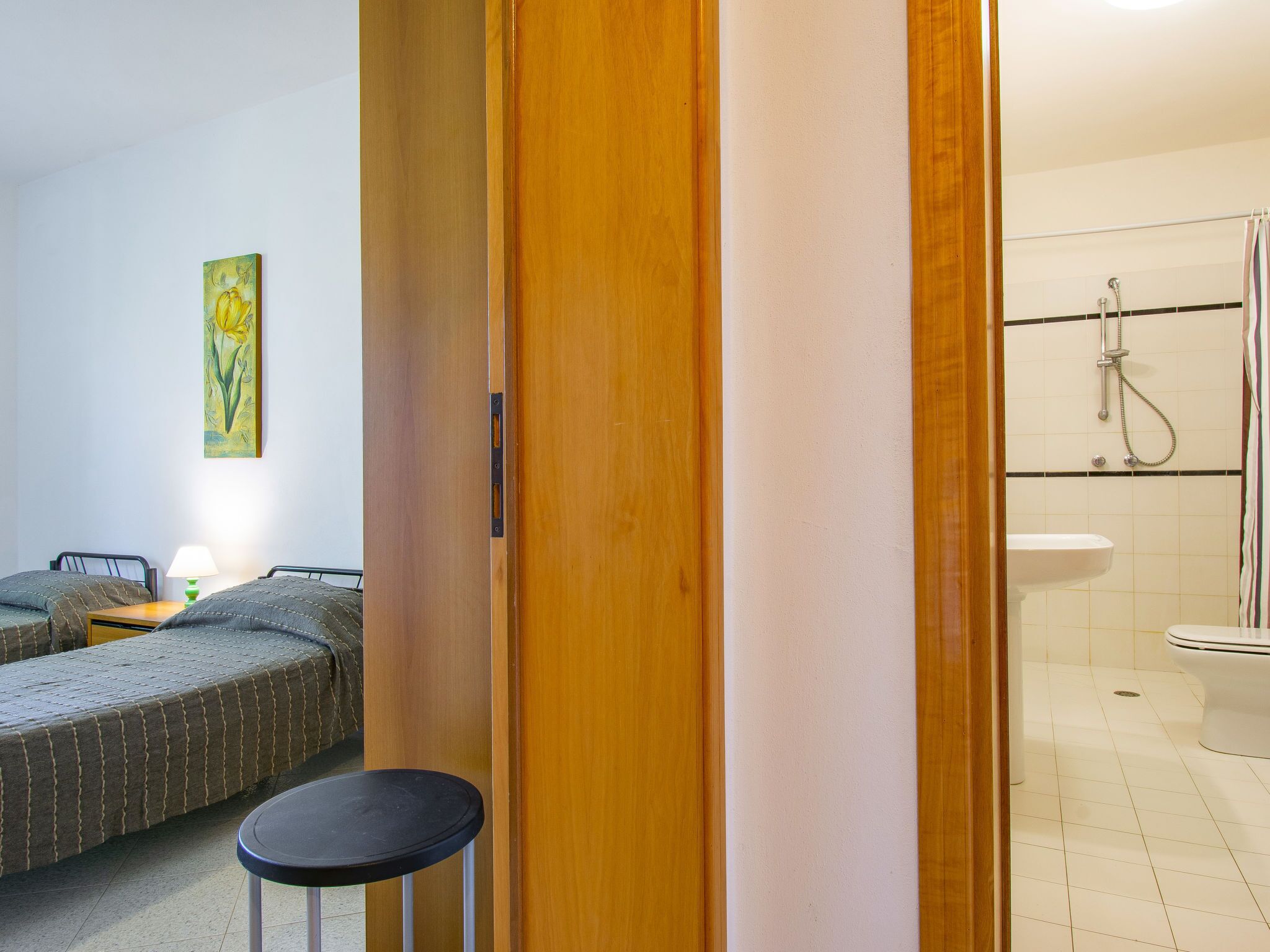 A hotel room with a wooden door, a bed with a green bedspread, a small round table, a painting on the wall, and a bathroom with a shower and sink.