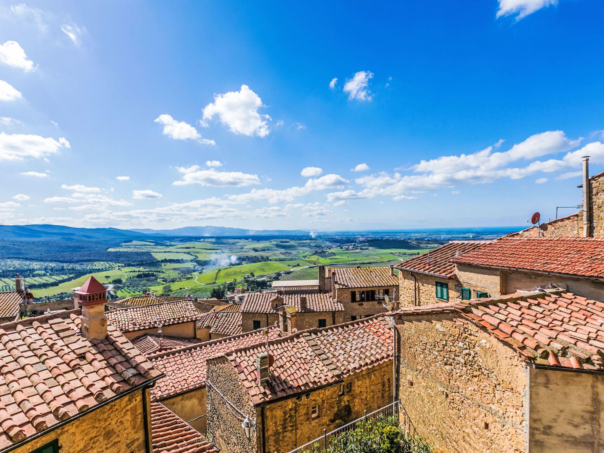 A view of a historic town with terracotta-roofed buildings, a clear blue sky, and rolling green hills in the distance.