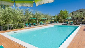 Ein rechteckiger Swimmingpool mit einer gefliesten Kante, umgeben von einer Pflasterterrasse. Mehrere grüne Sonnenschirme spenden Schatten, und einige Liegestühle sind sichtbar.