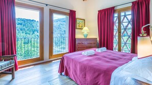 A bedroom with a large bed, a wooden floor, a nightstand with a lamp, a dresser, and a window with a view of a forested area. - Policleto by Interhome (San Polo in Chianti)