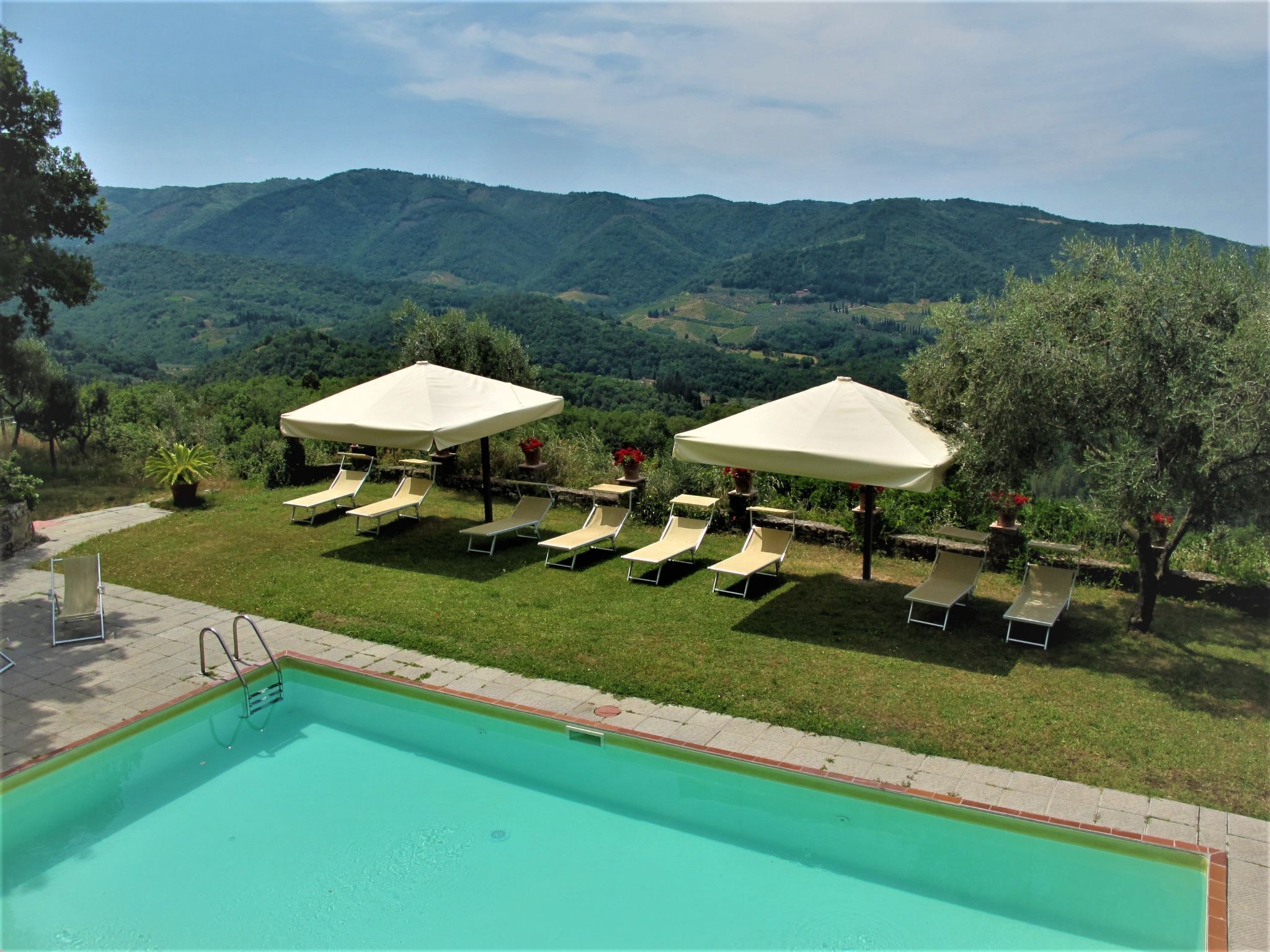 Zona de piscina con sillones de descanso y sombrillas, rodeada de exuberante vegetación y montañas.
