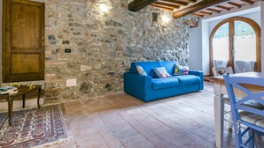 Ein rustikaler Raum mit Steinwand, Holzbalken, einem blauen Sofa, einer Holztür und einer Essecke mit Tisch und Stühlen.