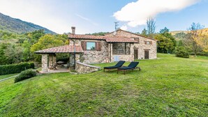 A stone house with a tiled roof, a patio with a table and chairs, and two lounge chairs in the yard.