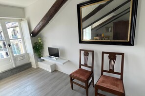 Una habitación con pared blanca, un espejo grande, un televisor sobre un estante blanco, dos sillas de madera y una planta en maceta.