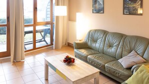 A living room with a green leather sofa, a wooden coffee table with fruit, a dining table outside, and two framed pictures on the wall. - Strandvilla (LUB117) by Interhome (Ostseebad Lubmin)