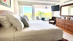 Une chambre avec un grand lit, un ventilateur de plafond, une télévision, une commode et un balcon offrant une vue sur les palmiers.