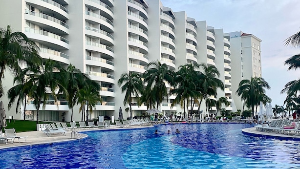 Une piscine dont le fond est recouvert de carreaux, entourée de palmiers et bordée d’une rangée d’immeubles à plusieurs étages dotés de balcons.