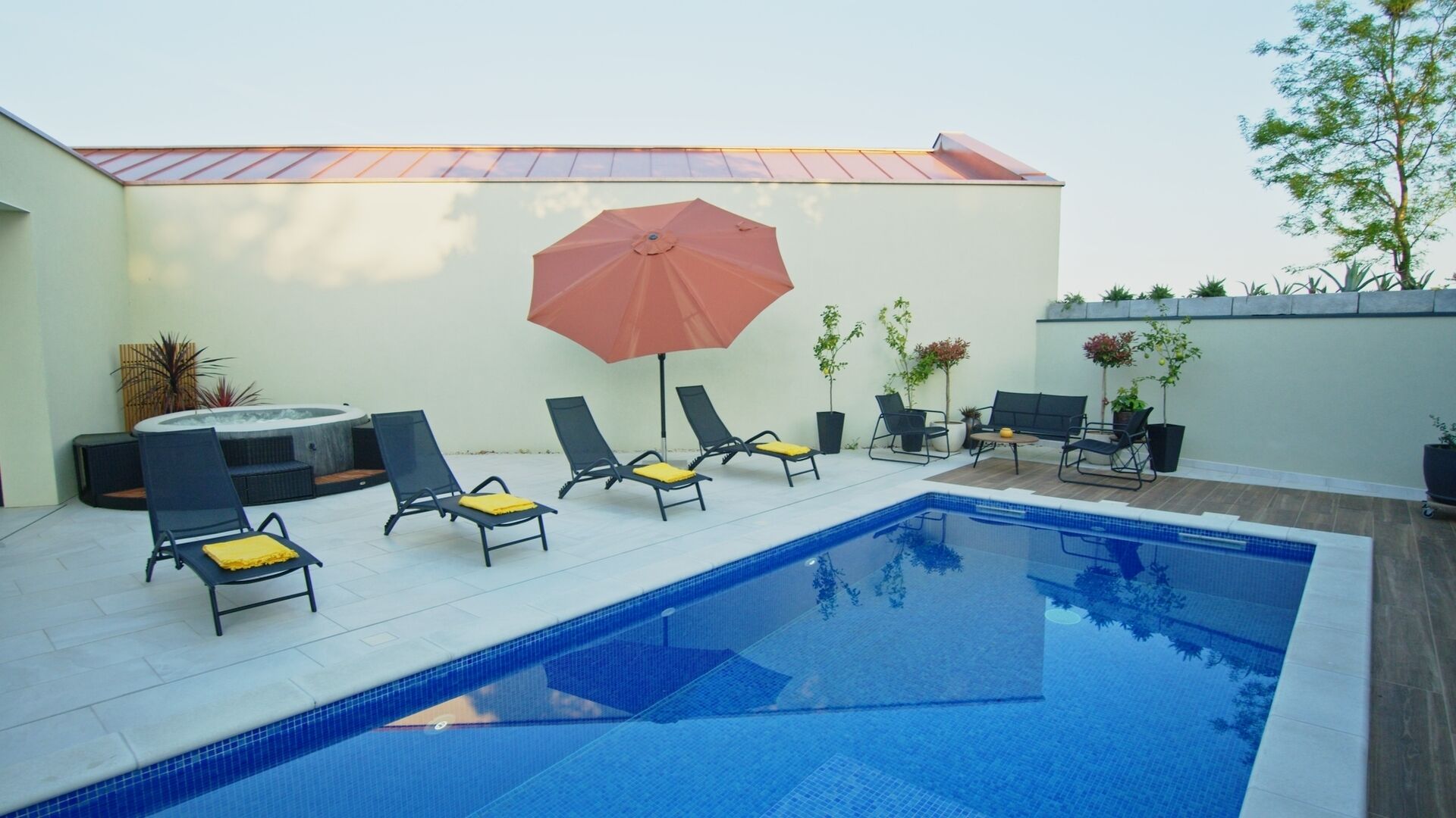 Une piscine entourée de plantes en pot, agrémentée de chaises longues et d’un parasol, ainsi qu’un jacuzzi.