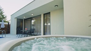 A modern outdoor hot tub area with a table and chairs, a wooden deck, and a glass-enclosed patio.