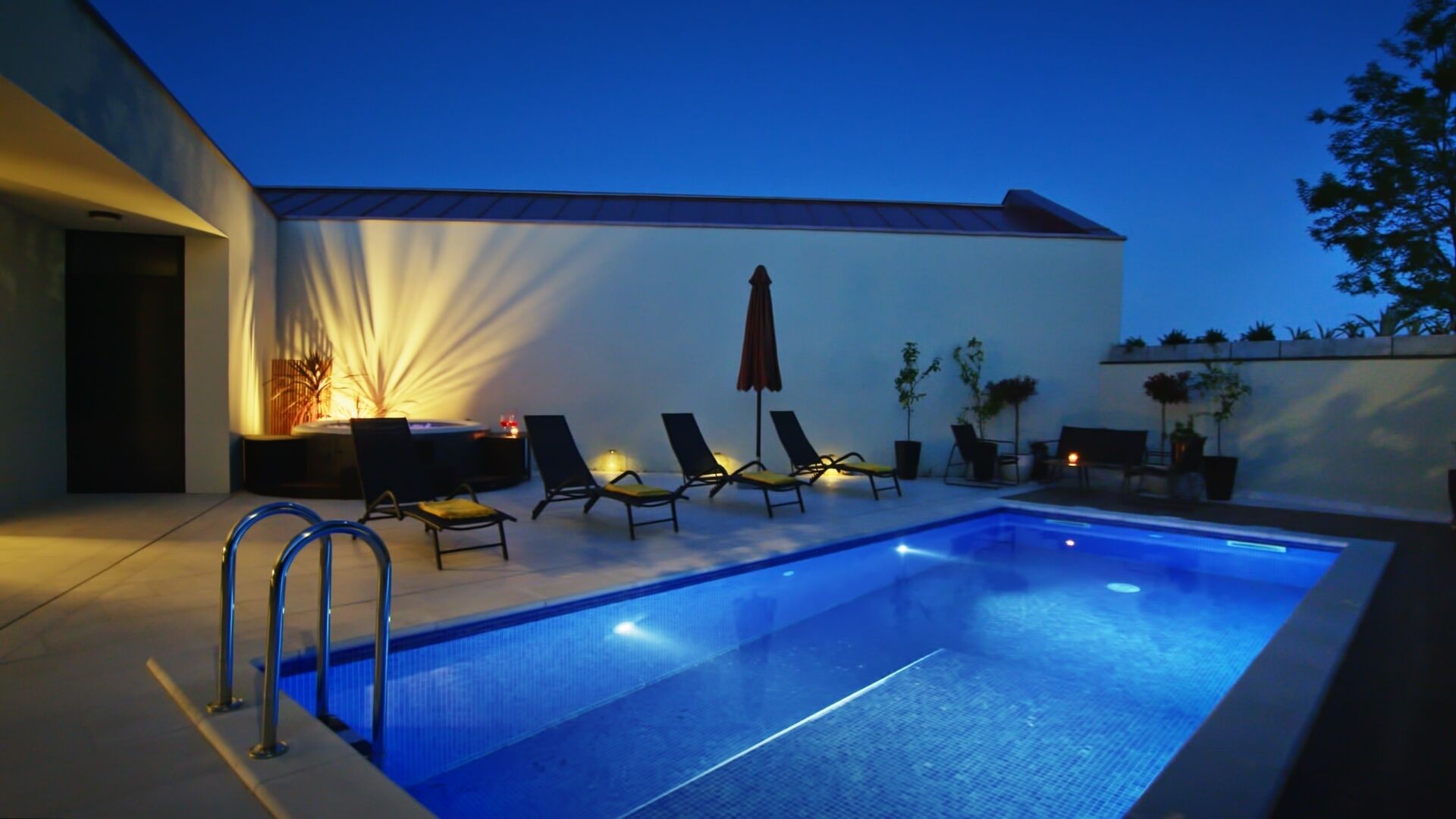 Piscina com espreguiçadeiras e uma mesa, cercada por um muro branco e plantas.