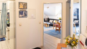 Un couloir mène à un espace de vie comprenant un canapé, une table basse et un fauteuil. Un miroir est accroché au mur, et une porte donne accès à une autre pièce.