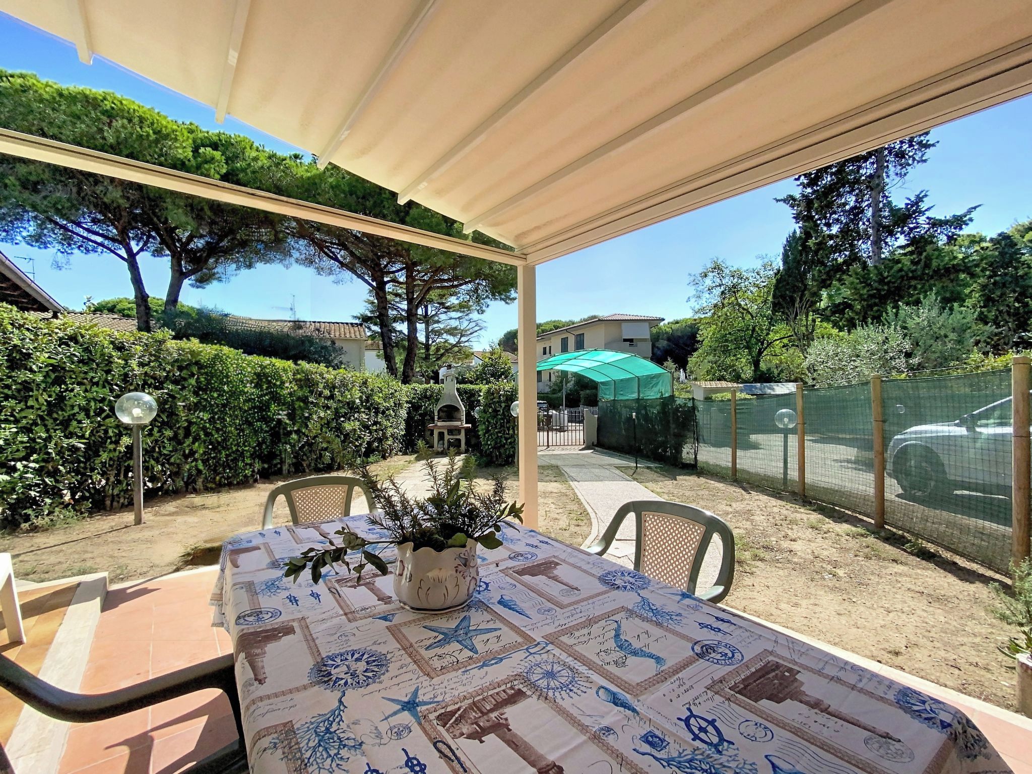 Un patio al aire libre con una mesa cubierta por un mantel con motivos náuticos, una planta en maceta y una sombrilla verde al fondo.