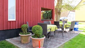 En rød bygning med et vindue og en dør, udendørs siddepladser med stole og et bord, samt plantekasser med planter.