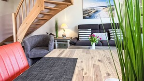 Ein modernes Wohnzimmer mit einer Wendeltreppe aus Holz, einem grauen Sofa, einem roten Sessel, einem Holztisch und einem Gemälde mit Strandmotiv.
