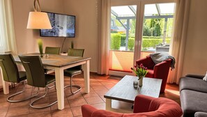 Ein modernes Wohnzimmer mit Essbereich, ausgestattet mit einem Fernseher, grünen Stühlen, einem roten Sofa und Blick durch große Glasüren.