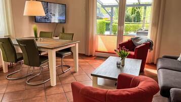 Ein modernes Wohnzimmer mit Essbereich, ausgestattet mit einem Fernseher, grünen Stühlen, einem roten Sofa und Blick durch große Glasüren.