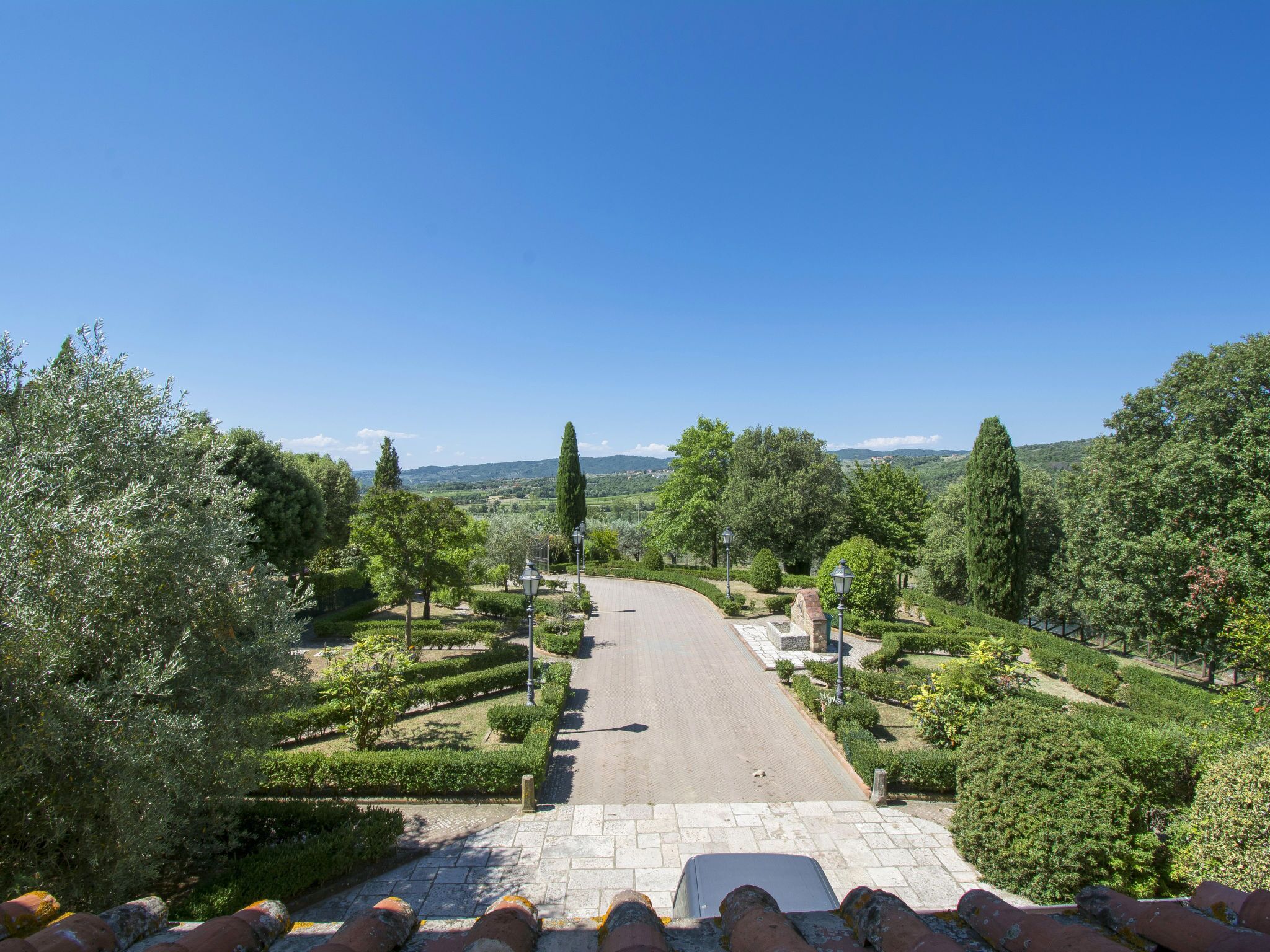 A paved pathway leading through a landscaped garden with neatly trimmed hedges and trees, under a clear blue sky.