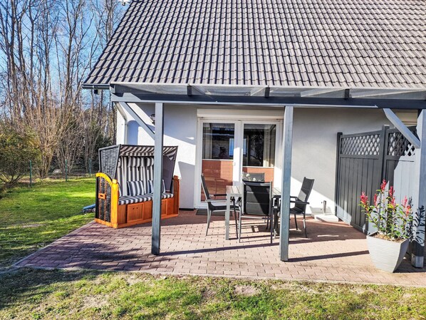 Una casa con techo de tejas, un porche cubierto con mesa y sillas, una terraza de madera y un jardín cercado.