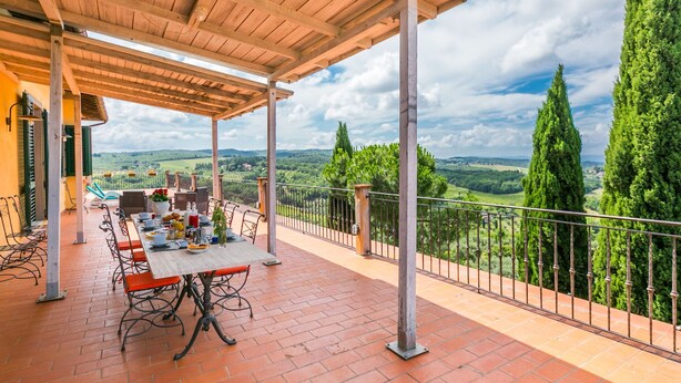 Un espace de restauration extérieur abrité, avec une table dressée pour quatre personnes, offrant une vue panoramique sur un paysage pittoresque de collines ondulantes et de grands arbres.