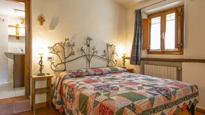 Une chambre à coucher avec un lit, deux tables de chevet, une fenêtre avec des rideaux, un radiateur, et une salle de bain visible au fond.