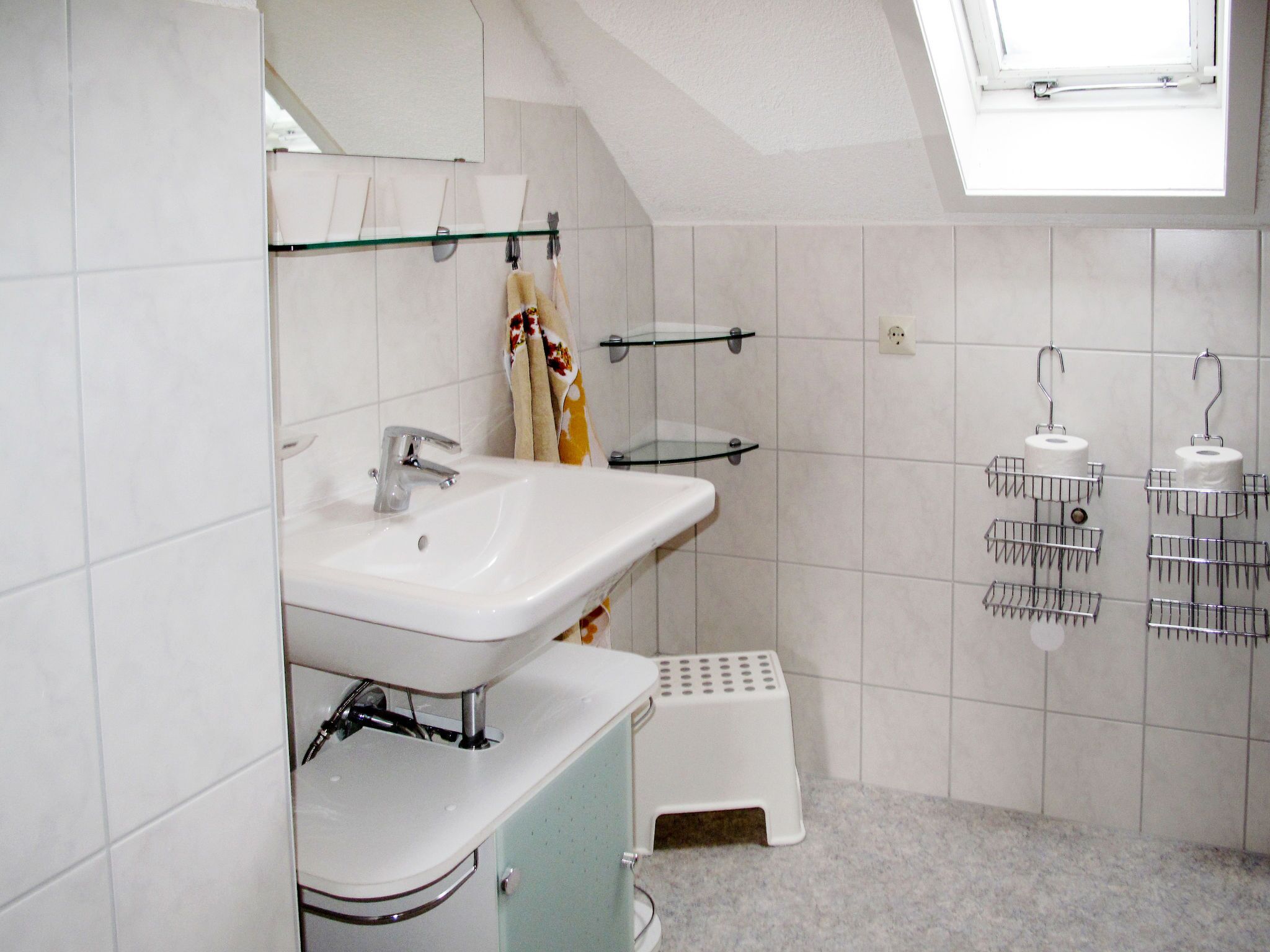 Baño con lavamanos blanco, espejo y zona de ducha. Hay dos repisas con artículos de aseo personal y un taburete pequeño.