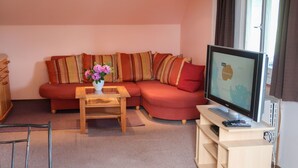 Un salon avec un canapé rouge, une table basse en bois sur laquelle est posé un vase de fleurs, un téléviseur sur un meuble et une petite armoire en bois.