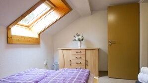 Une chambre avec un plafond en pente, un lit recouvert d’une couverture à carreaux violets, une commode en bois avec des tiroirs, une porte blanche et une petite fenêtre.