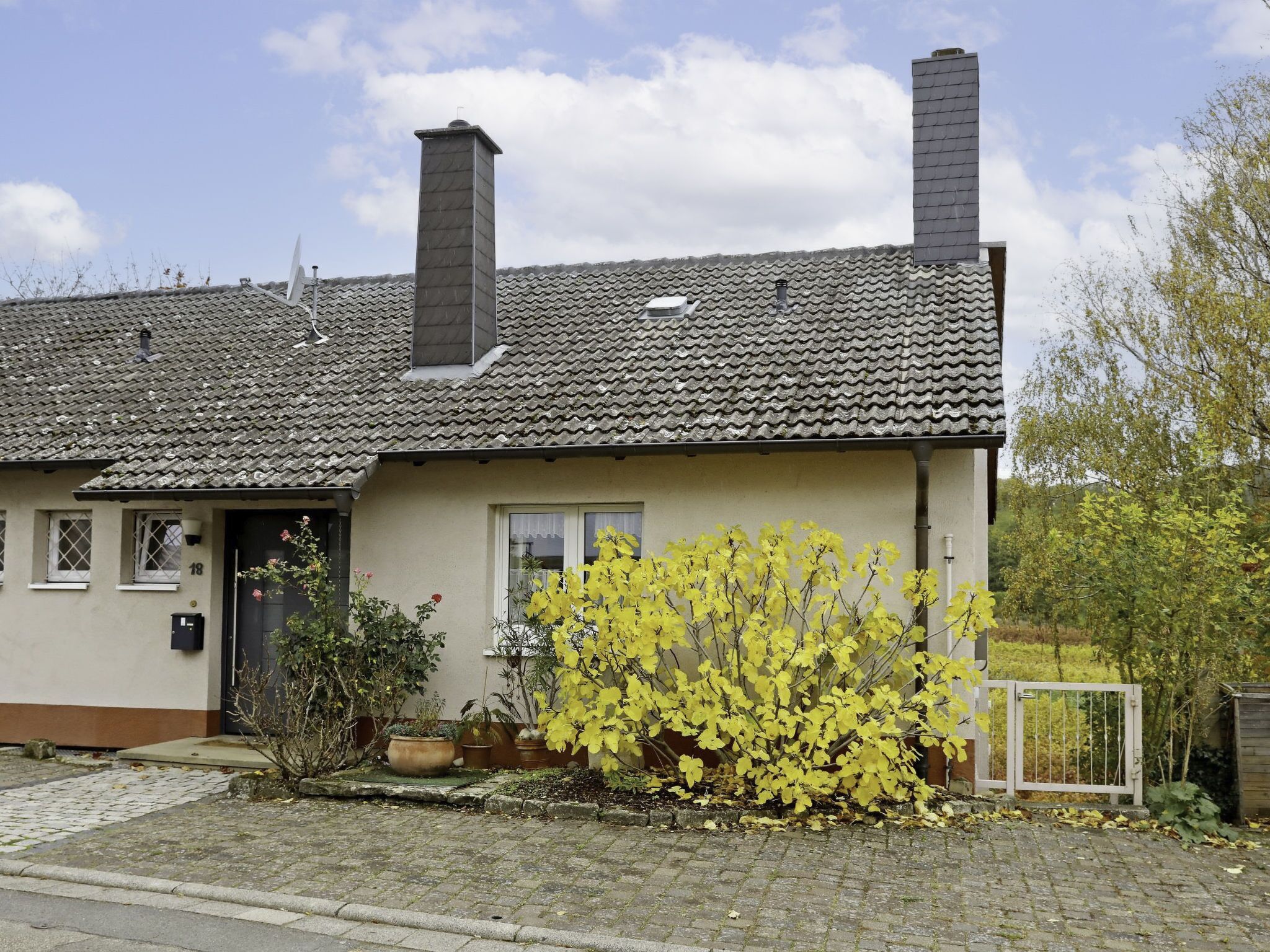 A two-story house with a tiled roof, a chimney, and a small garden with yellow flowering plants.