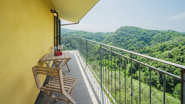 A balcony with a yellow wall, a table with chairs, and a view of a forested mountain range.