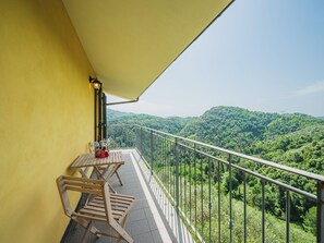 Een balkon met een gele muur, een tafel met stoelen en uitzicht op een bebost berglandschap.