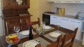 A dining area with a wooden table set for four, including plates, wine glasses, and a bottle of wine. There is a fruit bowl on the table, and a kitchen area with white cabinets and a stove in the background.