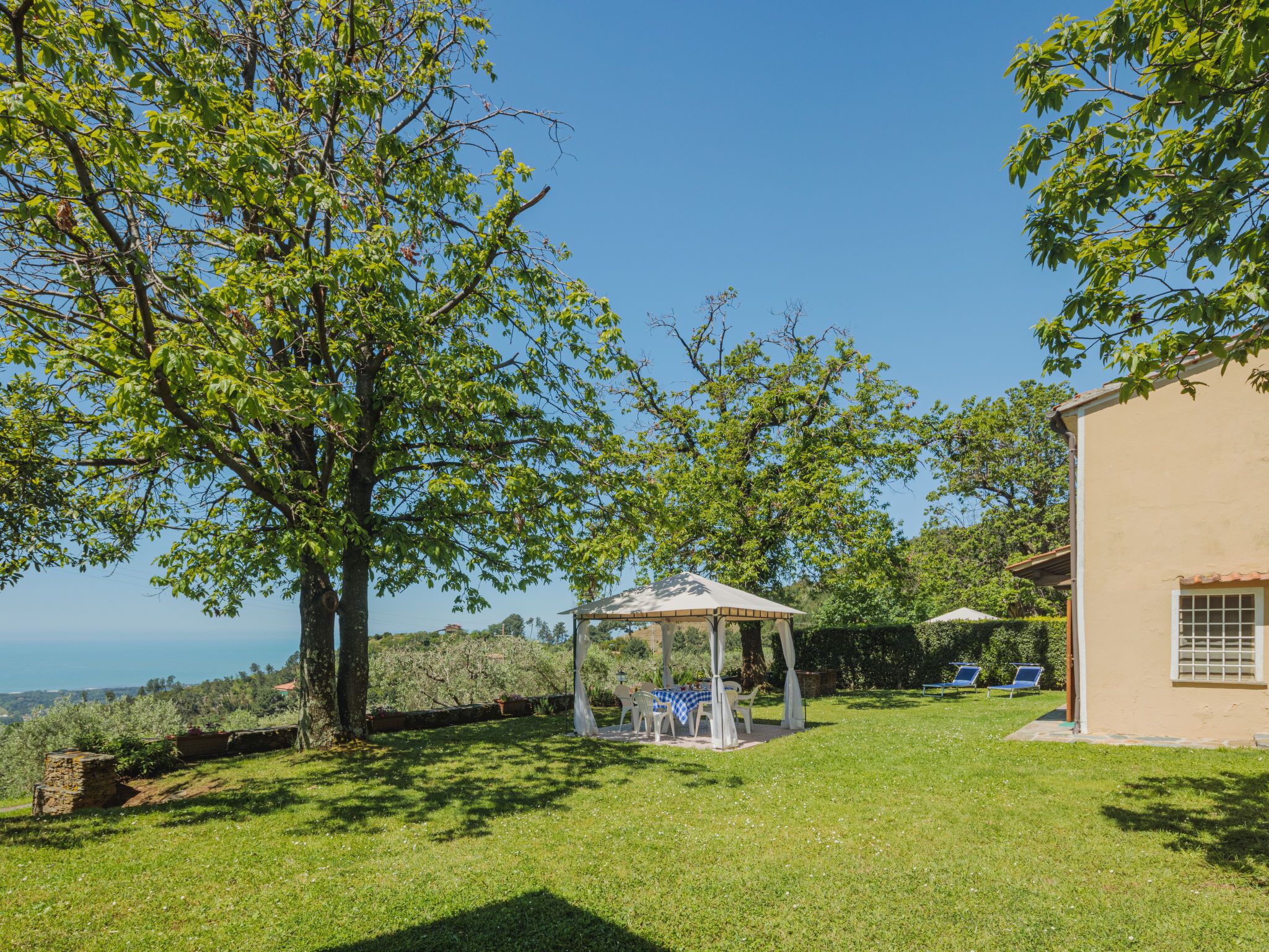 A sunny outdoor area with a gazebo, a table with chairs, and a building with a tiled roof.