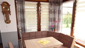 Un coin repas accueillant avec une table en bois et des chaises rembourrées, une horloge murale et une fenêtre avec des stores et un pot de fleurs.