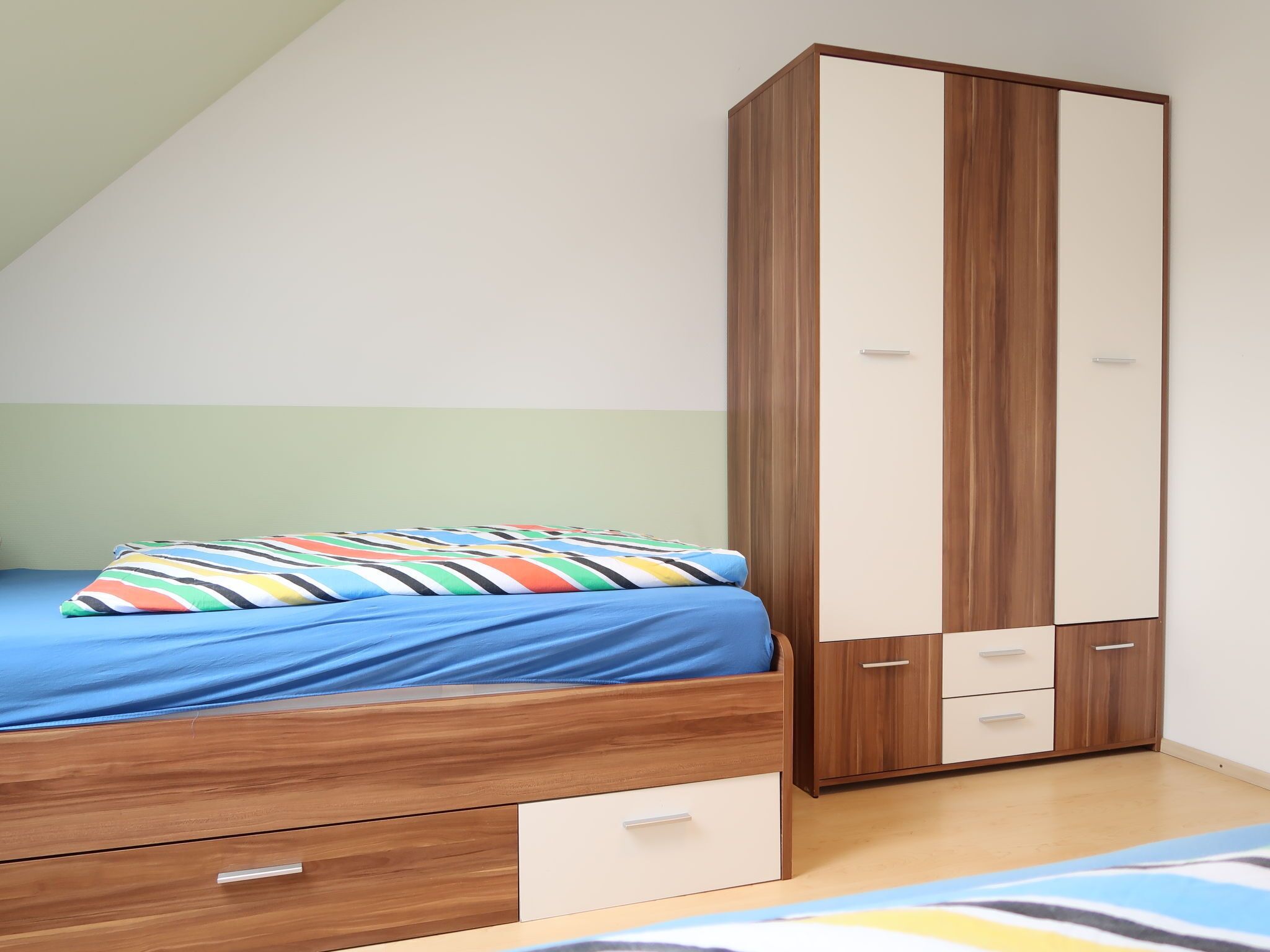 A bedroom with a bed, a wooden wardrobe, and a small drawer unit.