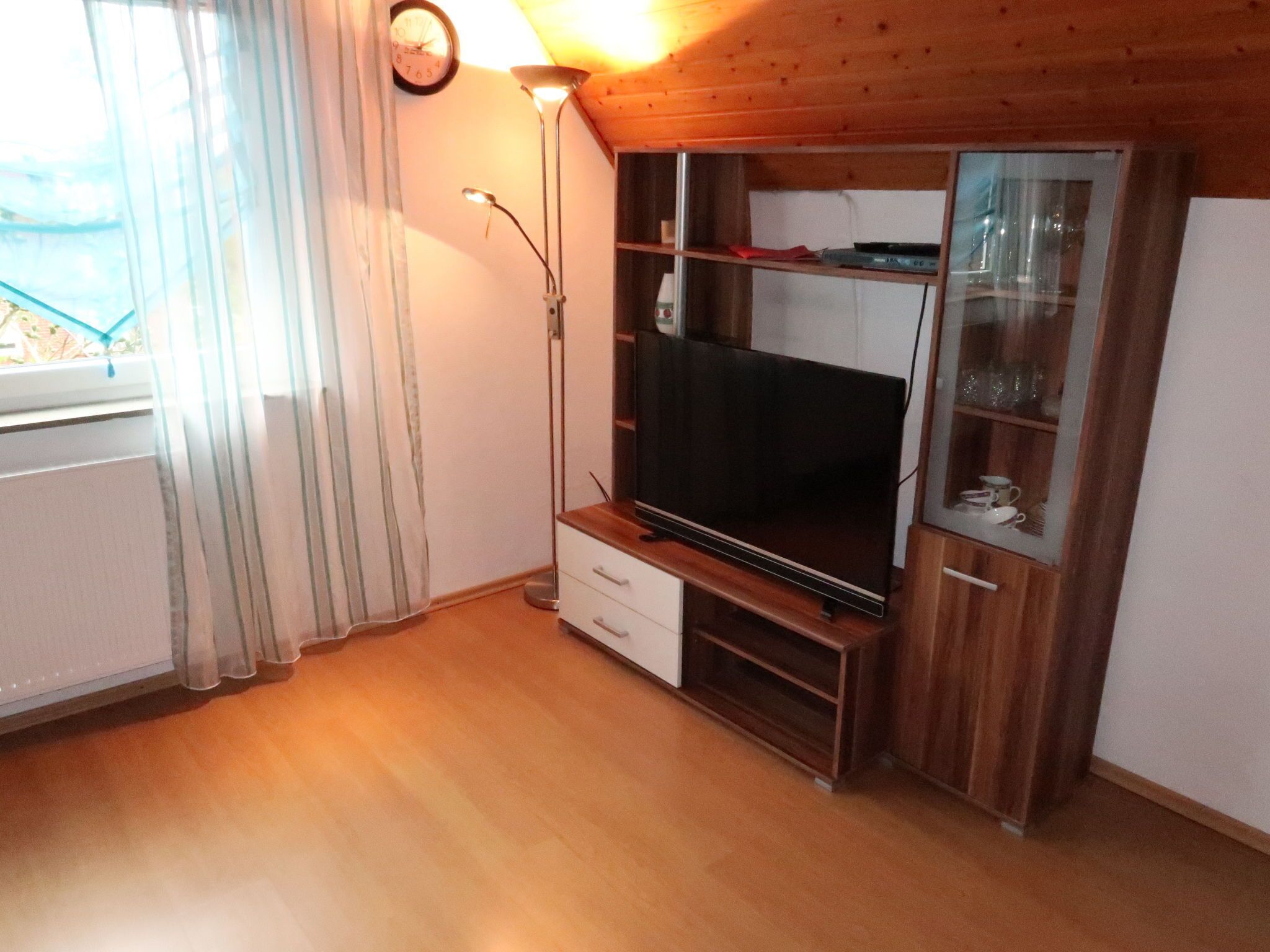 Une pièce avec un plancher en bois, un grand téléviseur à écran plat posé sur un meuble en bois, une vitrine avec des étagères, une horloge au mur et une fenêtre avec des rideaux.