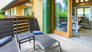 Un espacio al aire libre con sillas de metal y una mesa, un portón corredizo de vidrio que da a una cocina y una terraza de madera con una puerta verde.