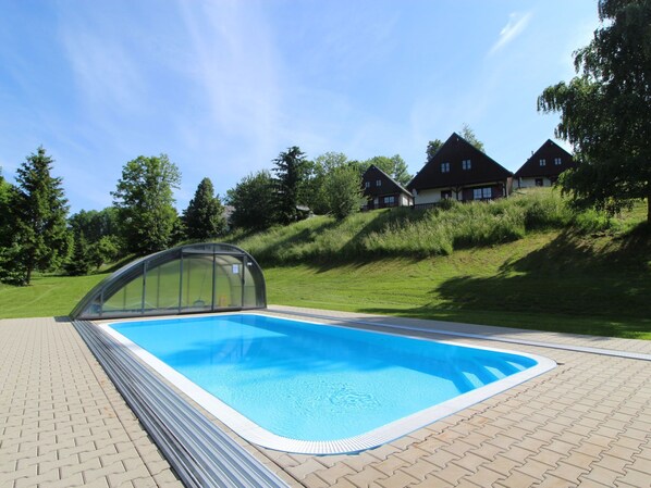 A swimming pool with a glass enclosure, surrounded by a paved area and a grassy hill with houses in the background.