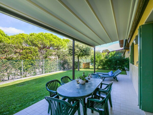 A covered patio with a table and chairs, a view of a garden, and a building with green doors.