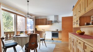 A modern kitchen with wooden cabinets, a dining area with a table and chairs, and a view of the outdoors through large windows. - Apartment Tirol by Interhome (Ortisei St Ulrich)